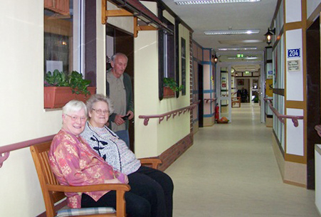Zwei ältere Frauen sitzen auf einer Bank im Gang eines Seniorenwerks.